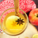 An overhead image of a warm hot toddy cocktail served with an apple slice and star anise garnish. A whole apple and lemon slices sit next to the glass.