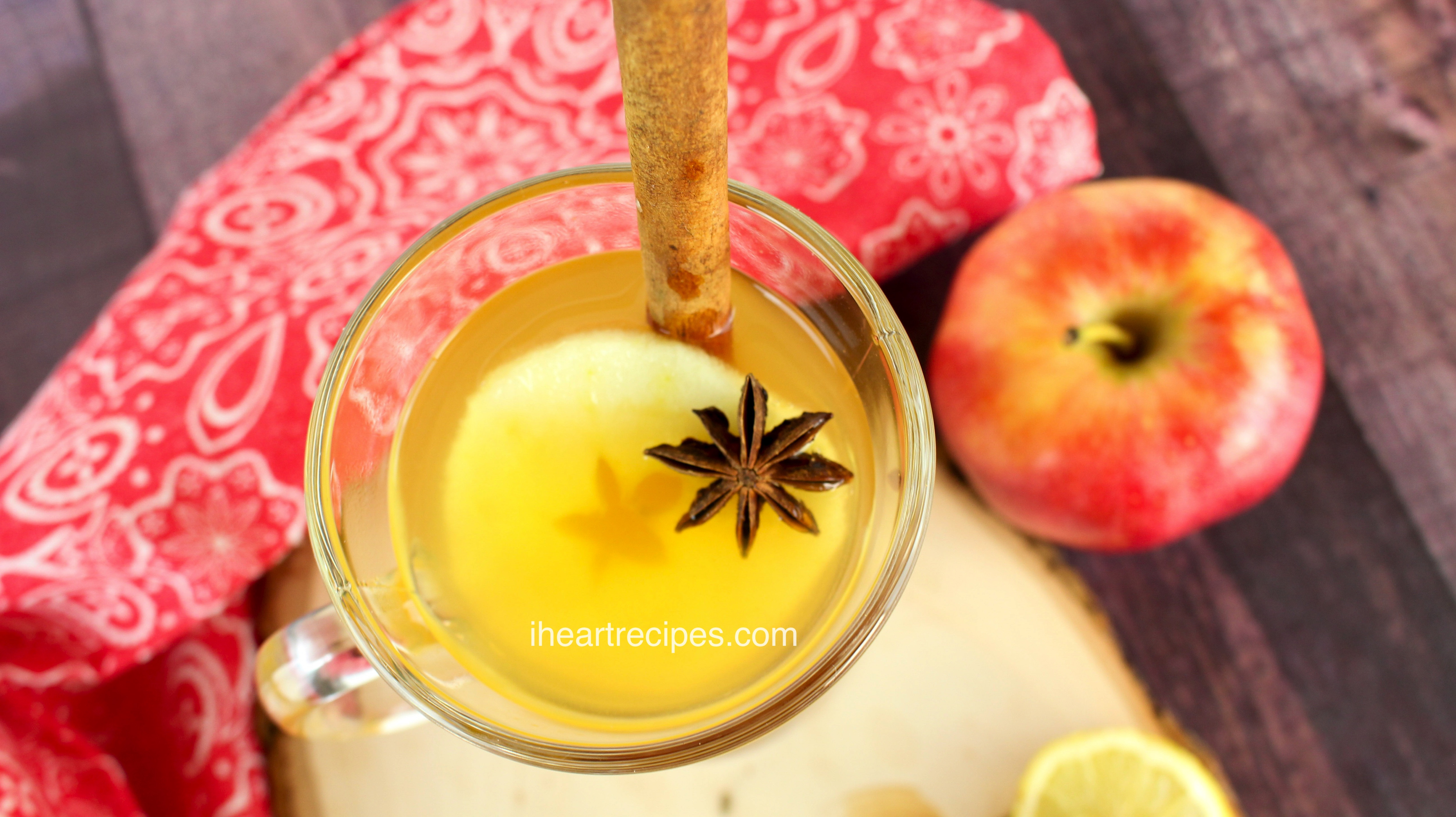 An overhead image of a warm hot toddy cocktail served with an apple slice and star anise garnish. A whole apple and lemon slices sit next to the glass.