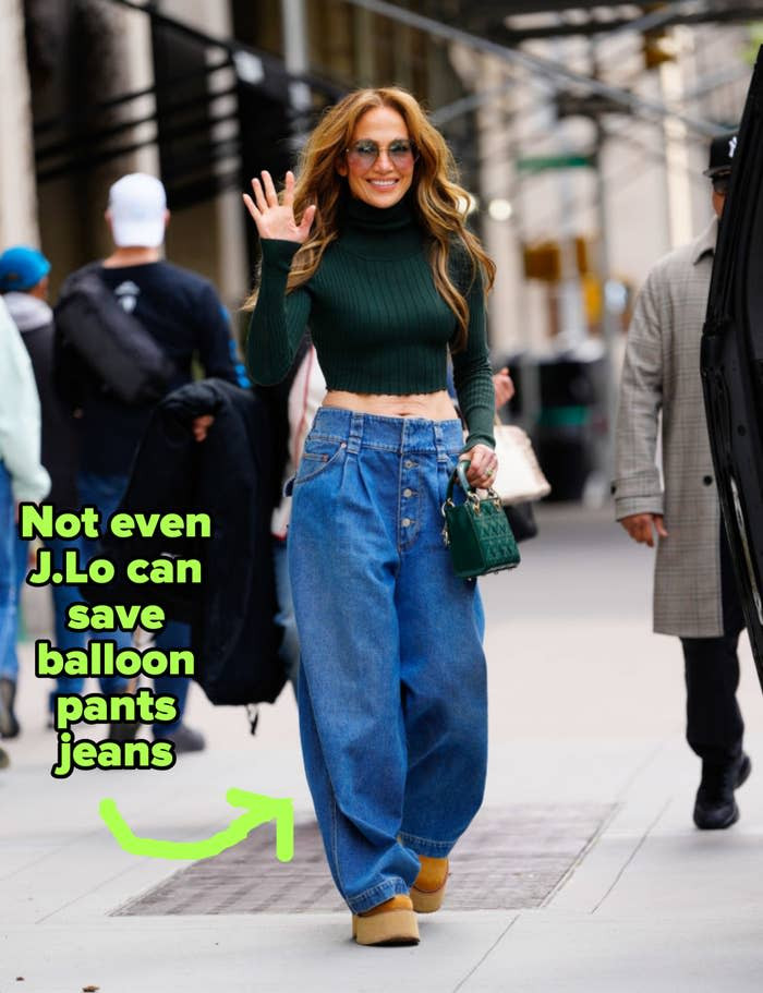 Person in a ribbed turtleneck crop top and wide-leg jeans, waving while walking on a city sidewalk.