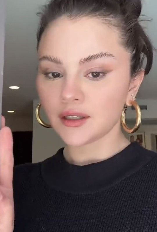 Close-up of a person with tied-back hair, large hoop earrings, and a black top facing the camera. On-screen app interface elements are visible.