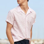 A group of stylish men are walking on a sandy beach, showcasing various beach outfits including linen shirts, polo shirts, swim shorts, and sunglasses, embodying the essence of men's beach fashion.