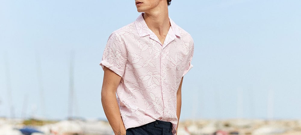 A group of stylish men are walking on a sandy beach, showcasing various beach outfits including linen shirts, polo shirts, swim shorts, and sunglasses, embodying the essence of men's beach fashion.