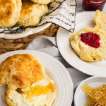 A basket of buttermilk biscuits on a woven mat, with two plates each featuring a biscuit; one with honey and butter, and the other with butter and red jam. Honeycomb and butter in background.