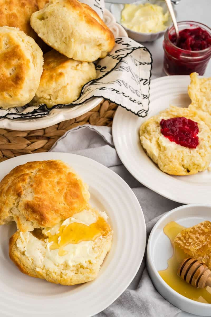 A basket of buttermilk biscuits on a woven mat, with two plates each featuring a biscuit; one with honey and butter, and the other with butter and red jam. Honeycomb and butter in background.