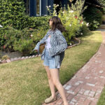 Woman wearing a red striped button down shirt, white tank top, denim shorts, brown clogs, and carrying a raffia tote bag.