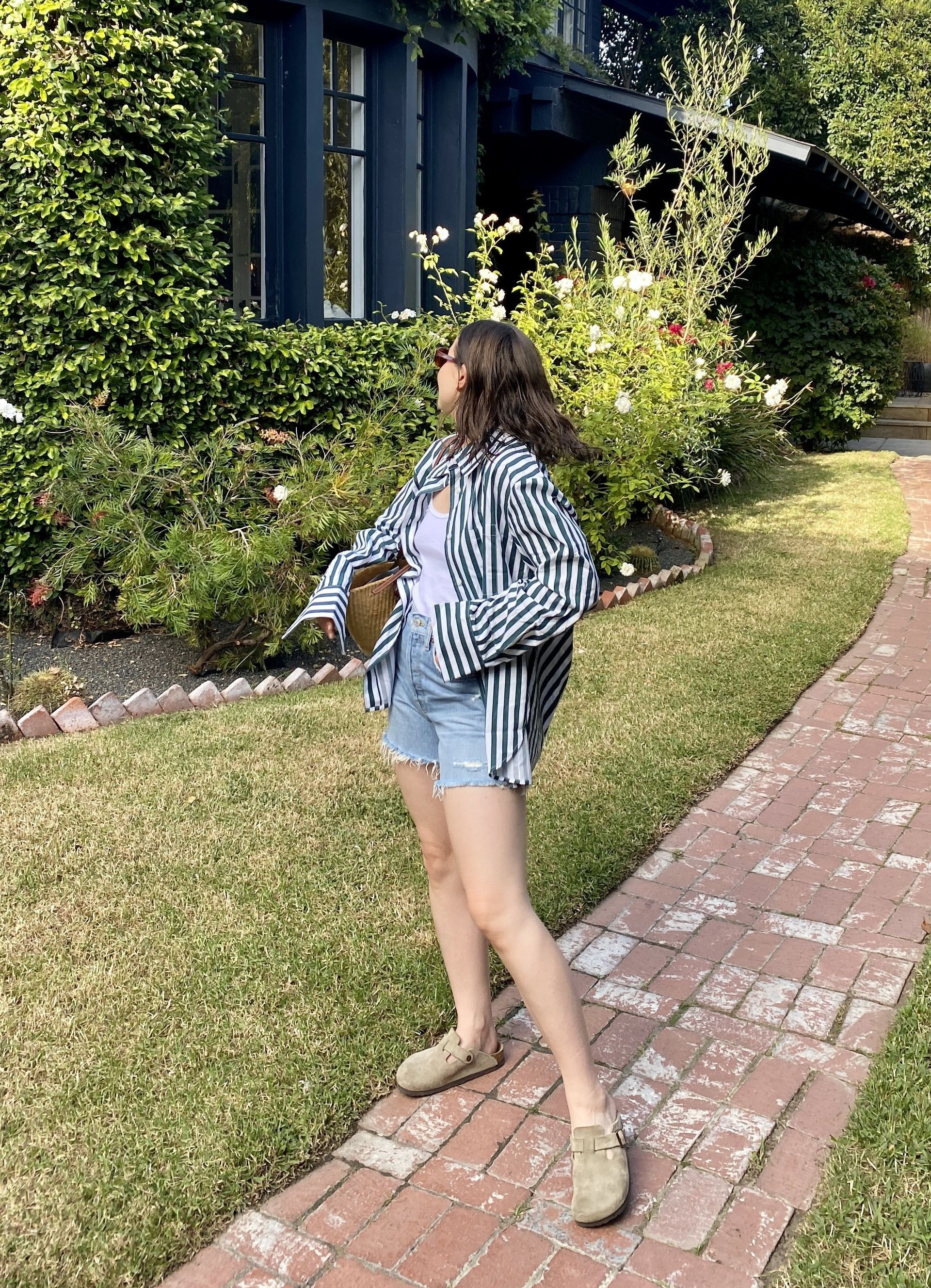 Woman wearing a red striped button down shirt, white tank top, denim shorts, brown clogs, and carrying a raffia tote bag.