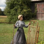 Woman in pioneer clothing standing in a field wearing a long-sleeved blue top and long blue skirt.