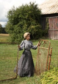 Woman in pioneer clothing standing in a field wearing a long-sleeved blue top and long blue skirt.