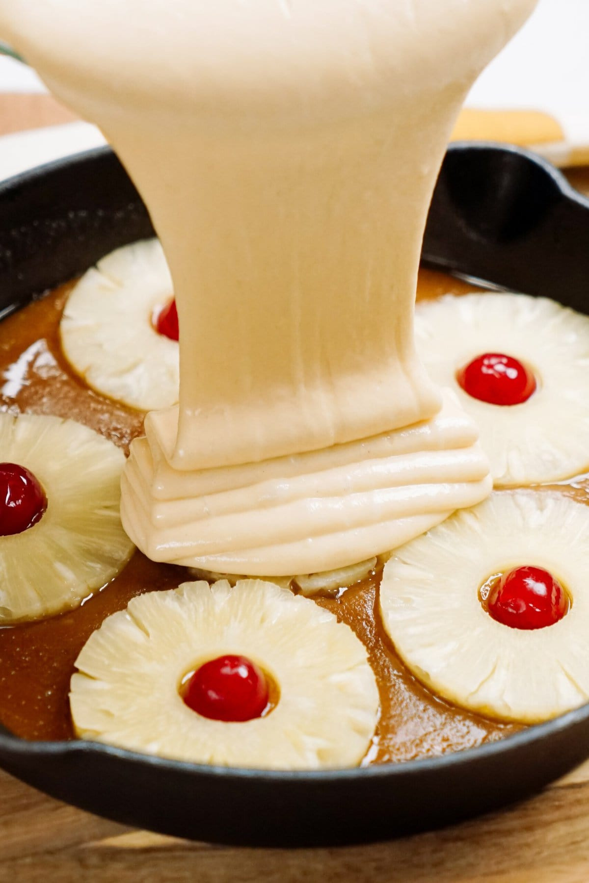 Pouring cake batter over pineapple slices and cherries in a pan, a key step in making a delicious upside-down cake.
