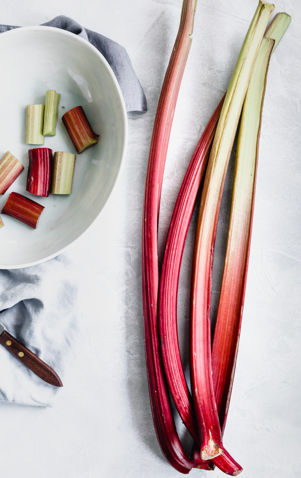 long stalks of fresh rhubarb next to a small knife and a bowl with rhubarb pieces