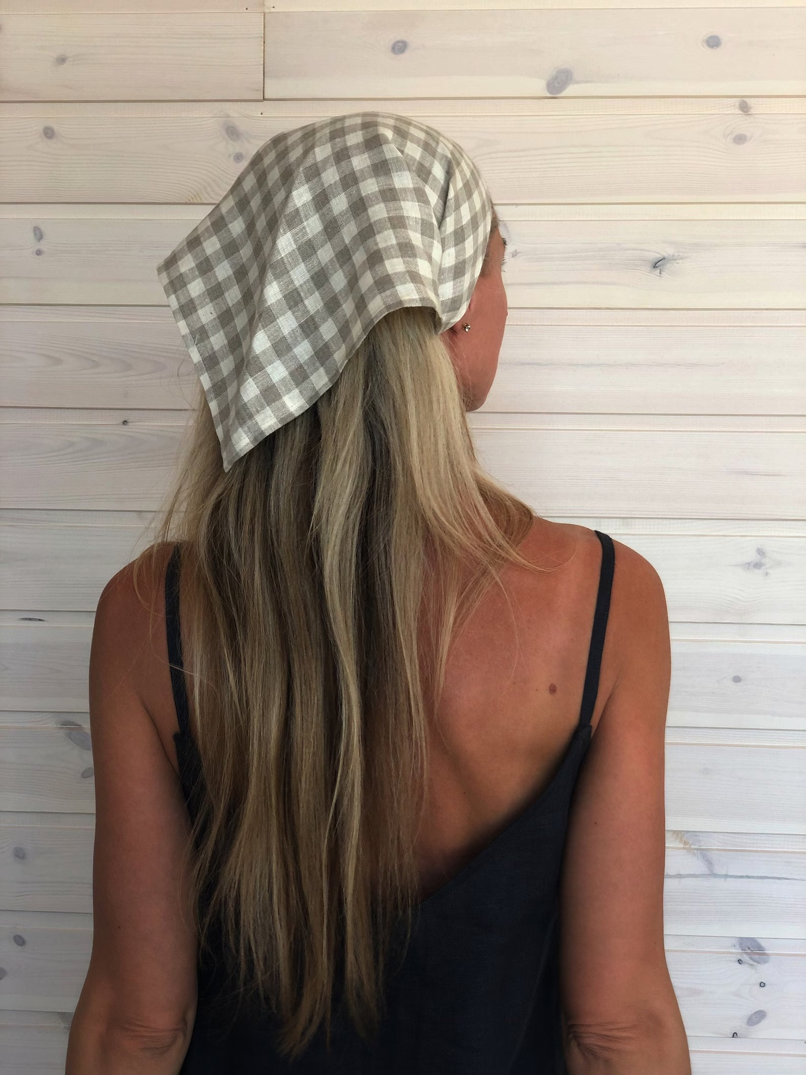 A person with long blonde hair wears a checkered headscarf and a black camisole, standing with their back to the camera against a light wood-paneled wall, showcasing a vintage-inspired cottagecore style