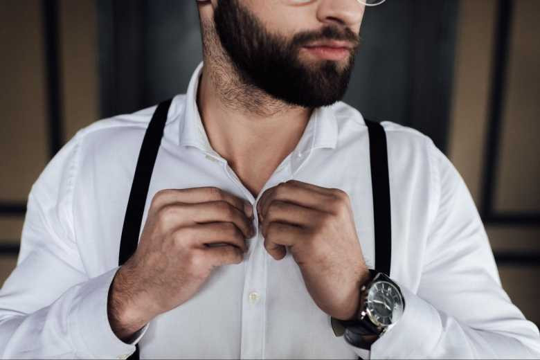 Man with beard buttoning up shirt while wearing suspenders