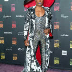 Billy Porter wears a huge wide brim hat in black and red, with a silver velvet wide collar lapel and a rhinestone jumpsuit.