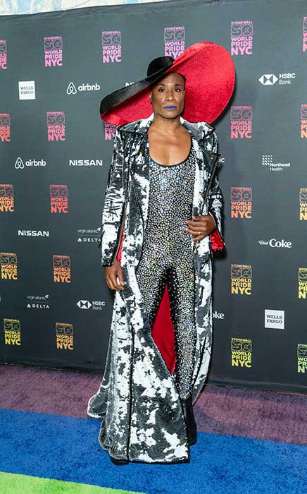 Billy Porter wears a huge wide brim hat in black and red, with a silver velvet wide collar lapel and a rhinestone jumpsuit.