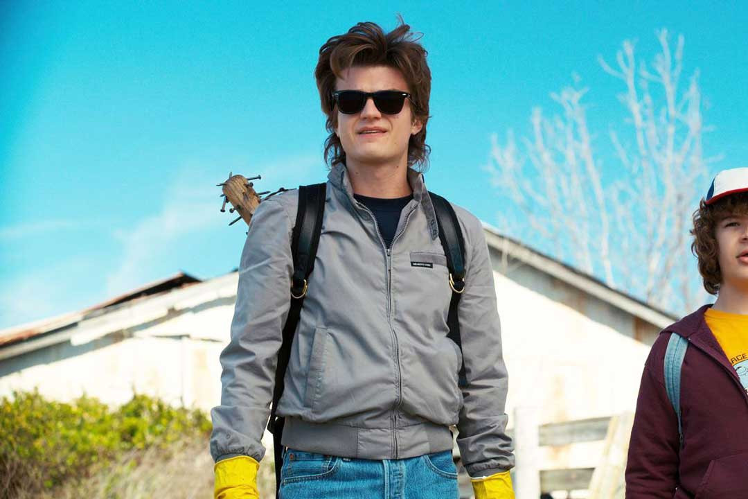 Character Steve Harrington from Netflix series Stranger Things wearing dark RayBan Wayfarer sunglasses, blue jeans and yellow rubber gloves