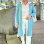 Woman over 60 wearing an organic linen cardigan, washable ivory silk blouse, tile white jeans, and alabaster woven tote, demonstrating soft white tones