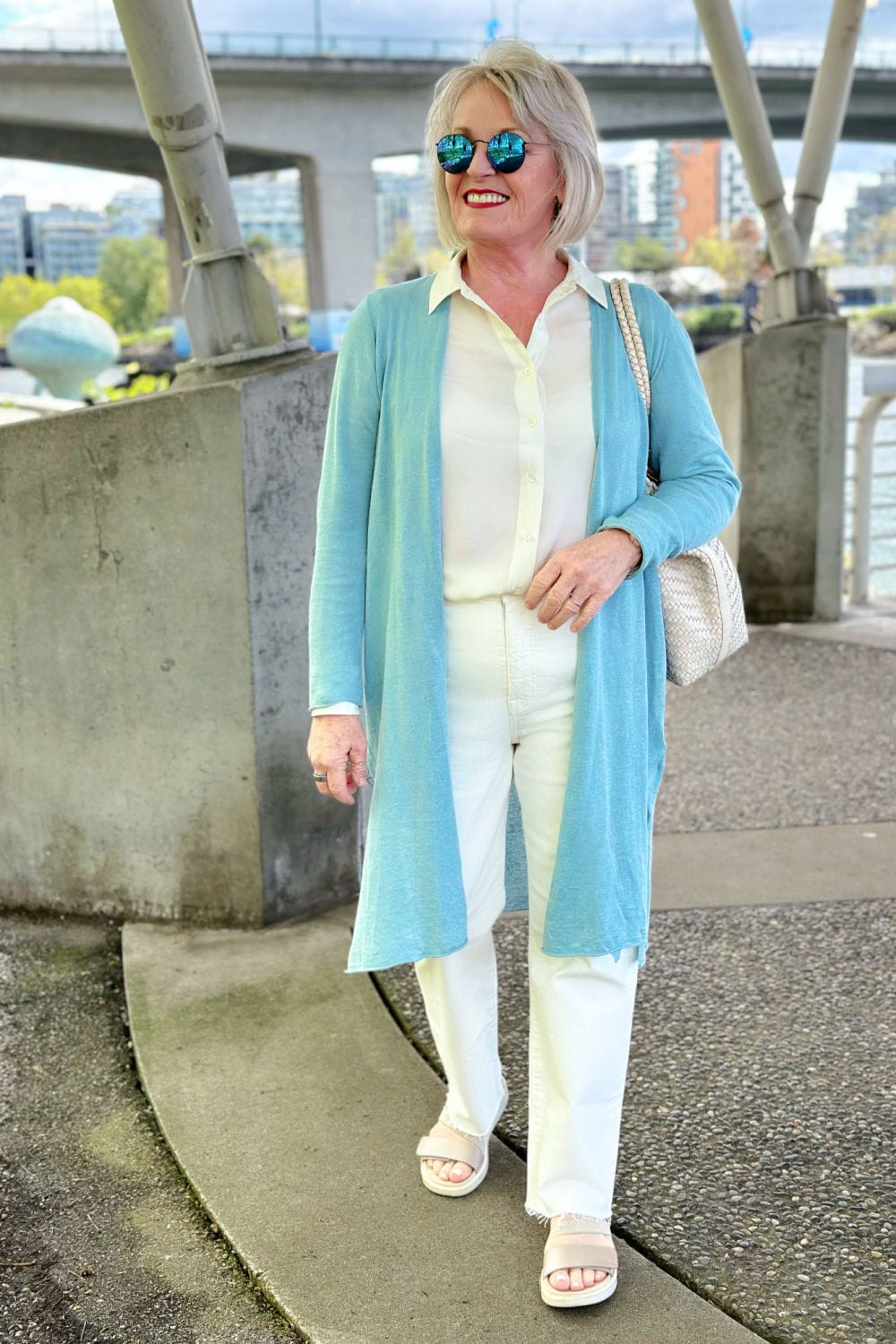 Woman over 60 wearing an organic linen cardigan, washable ivory silk blouse, tile white jeans, and alabaster woven tote, demonstrating soft white tones