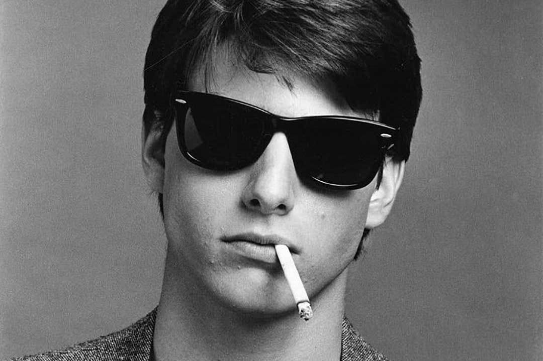 Greyscale image of Tm Cruise wearing black 80s style sunglasses with cigarette hanging out his mouth