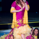 Jenni Rivera at Teleton 2010, wearing a fuchsia and gold dress, smiling brightly for the camera, showcasing her vibrant style.