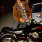 Pictured circa 1991, Keanu Reeves wears a shearling jacket with a plaid button-down as he gets on his motorcycle.