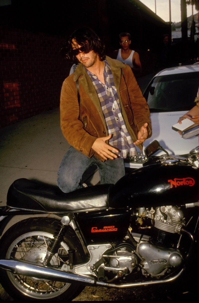 Pictured circa 1991, Keanu Reeves wears a shearling jacket with a plaid button-down as he gets on his motorcycle.