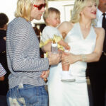 Kurt Cobain, Frances Bean Cobain, and Courtney Love attend the 1993 MTV Video Music Awards. Kurt wears a long-sleeve striped tee with distressed denim jeans and red sunglasses.