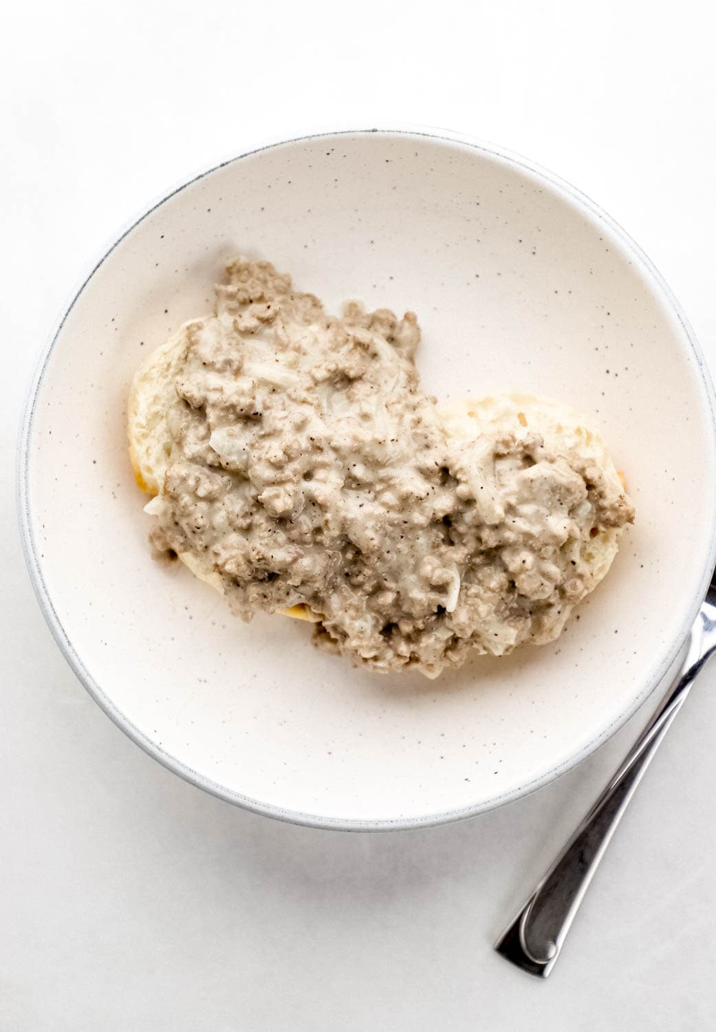 Overhead view of gravy on top of biscuit in white bowl by a fork on grey surface.