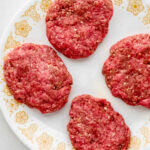 Four raw hamburger patties on white decorated plate.