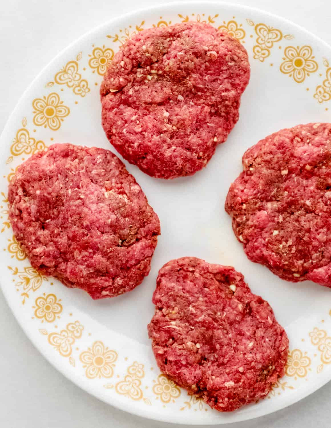 Four raw hamburger patties on white decorated plate.