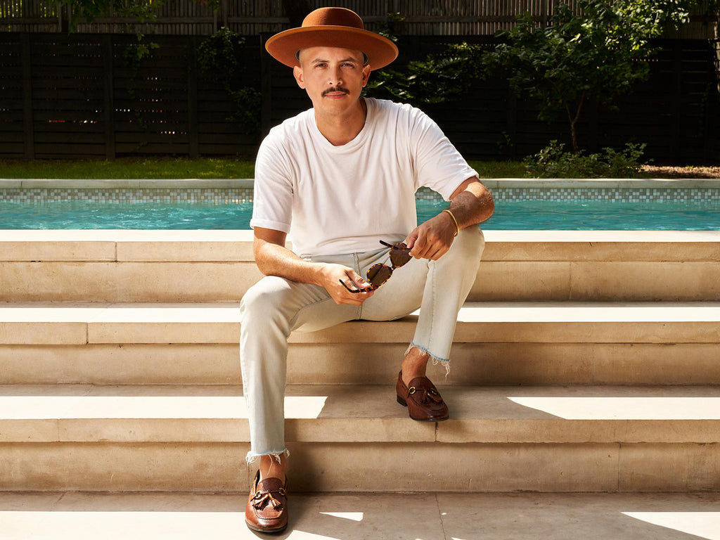 Man with a dark pencil mustache is wearing a brown fedora and sitting on the steps in front of a pool