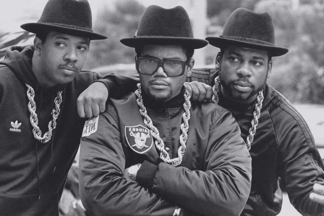 Run DMC rap group together wearing gold chains, hats and thick rimmed eyeglasses