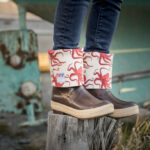 Photo of person wearing brown XTRATUF boots with the tops rolled down, featuring a red Octopus Salmon Sisters design. Person is standing on top of tree stump in front of an old, grounded fishing boat