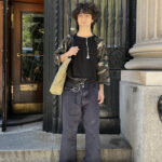 Student stands in front of a school building on Fifth Avenue wearing a raglan top with camouflage sleeves, black jeans, brown shoes, and a green tote bag