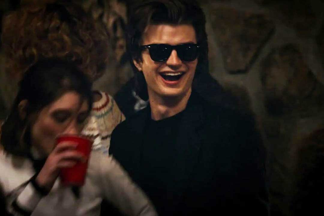 Steve Harrington from Netflix series Stranger Things wearing black wayfarer sunglasses at a party smiling
