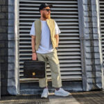 Man wearing a tan vest over a white t-shirt with chinos and sneakers, carrying a black briefcase