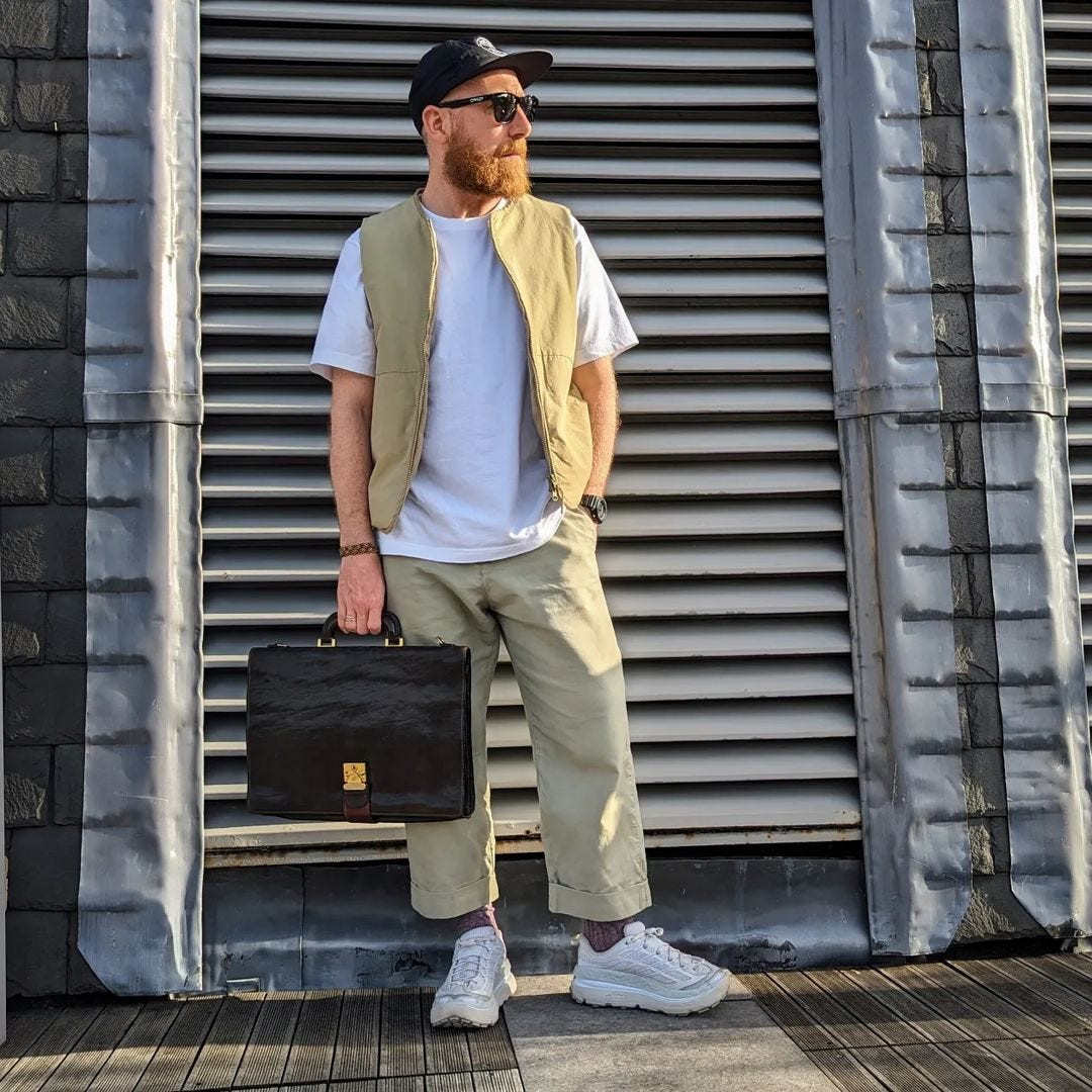 Man wearing a tan vest over a white t-shirt with chinos and sneakers, carrying a black briefcase