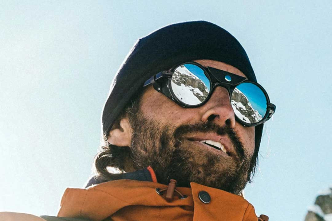 Upward view of man wearing hat jacket and round glacier sunglasses in the mountains
